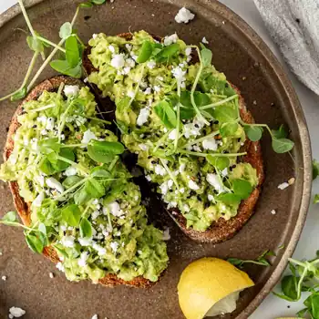 Two slices of avocado toast with herbs and a lemon wedge